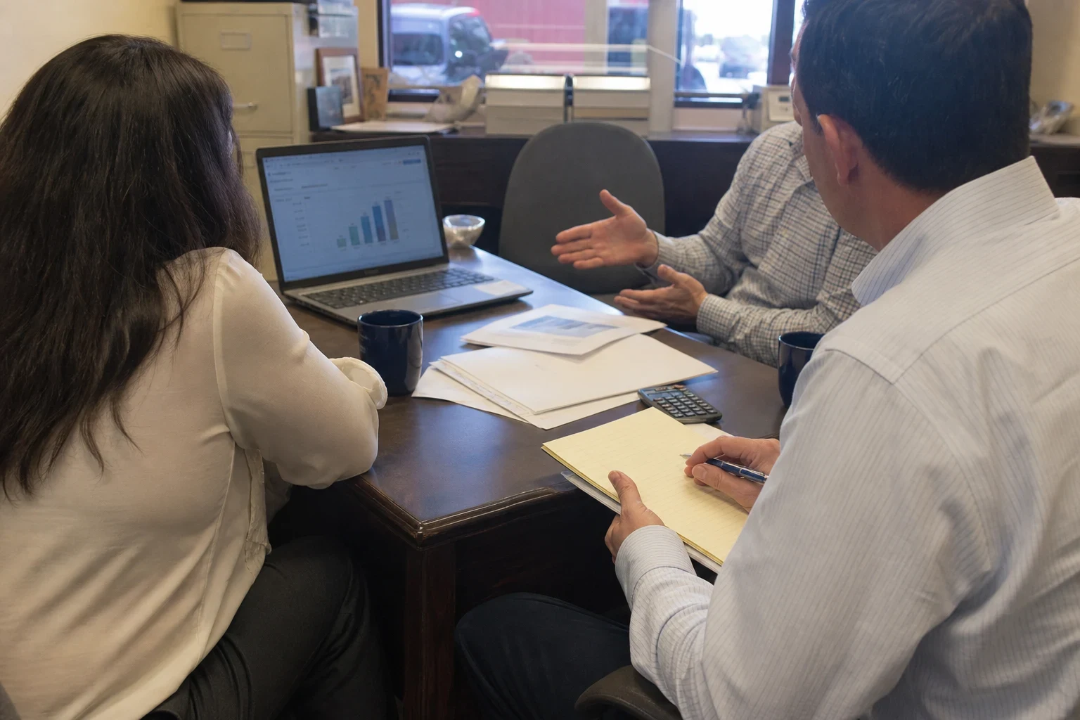Agent in a striped button-down takes notes on a legal pad while a man and woman review a bar chart on a laptop at a wooden table in a small storefront office