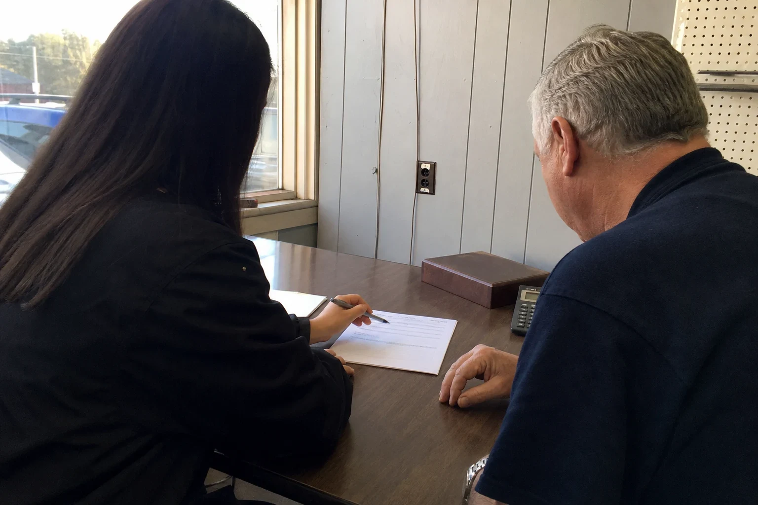 An agent with long dark hair points a pen at a short form on a wooden desk while a gray-haired man in a navy shirt reviews it in a small back office with paneled walls.